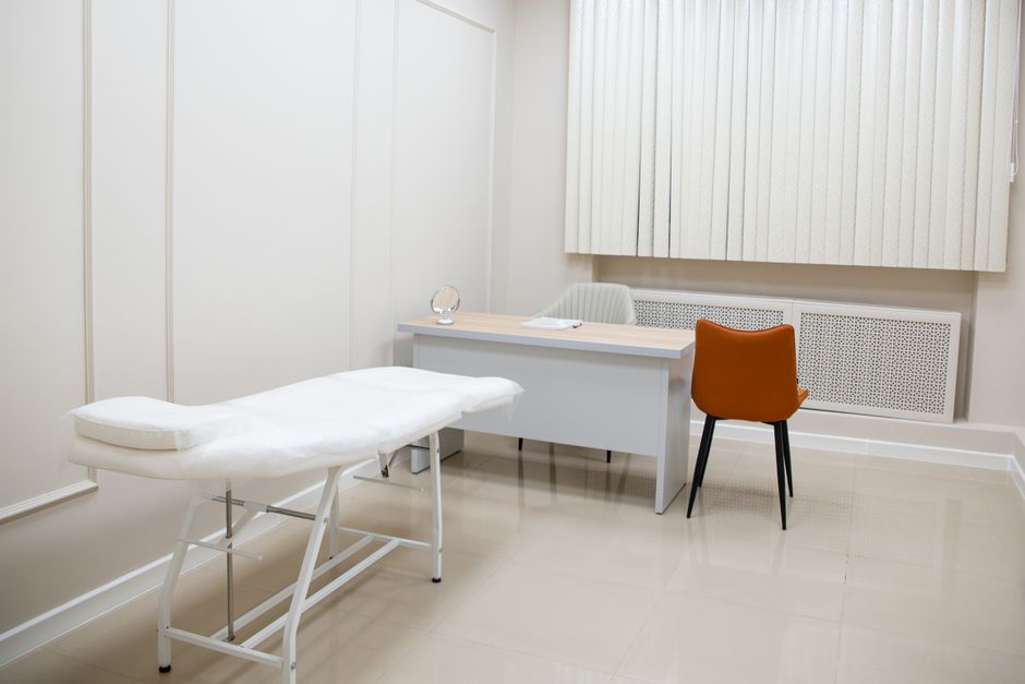 Consultorio médico con camilla de revisión y escritorio de madera junto a la ventana